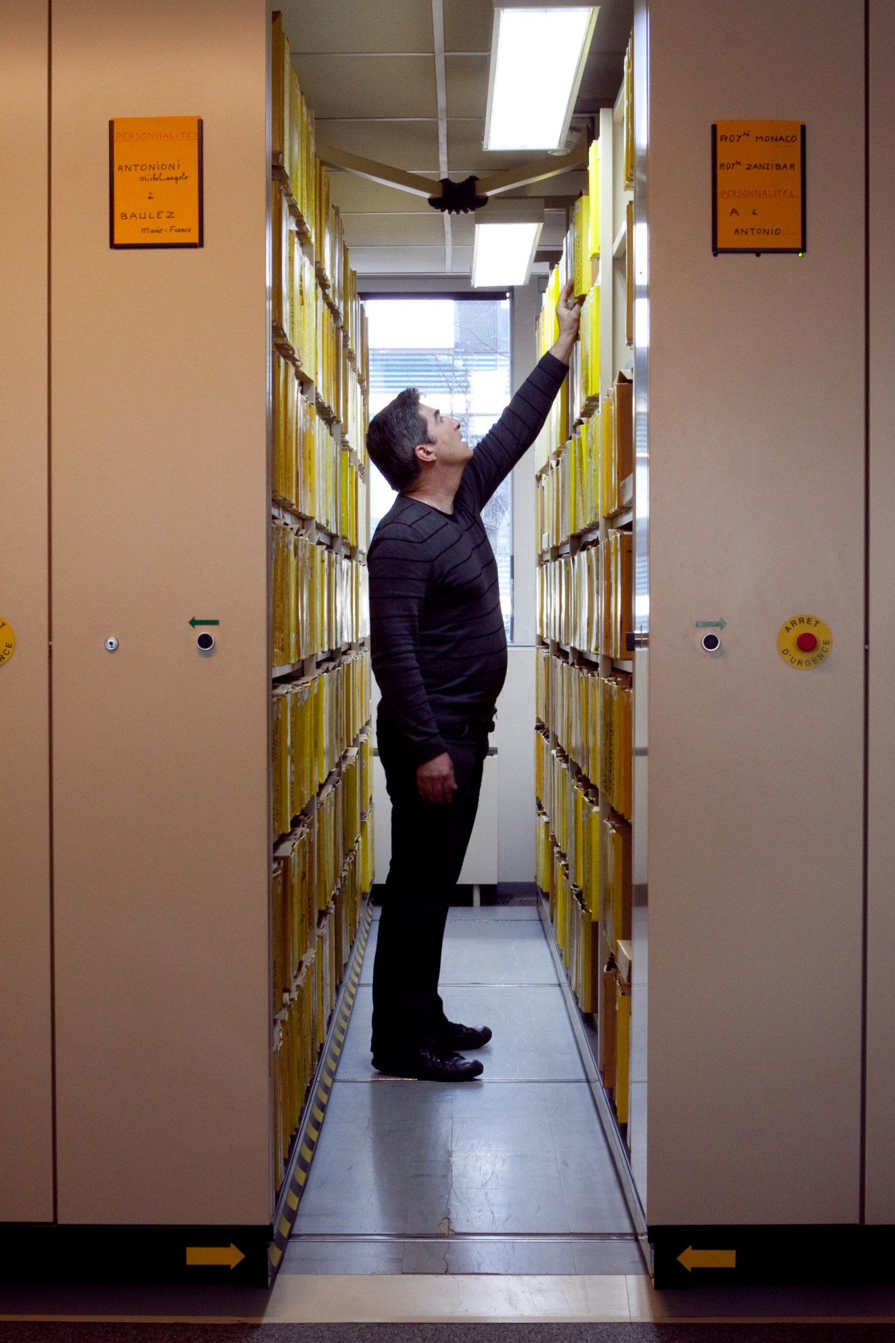 A man reaches up inside a narrow corridor lined with yellow filing cabinets containing archived photographs.