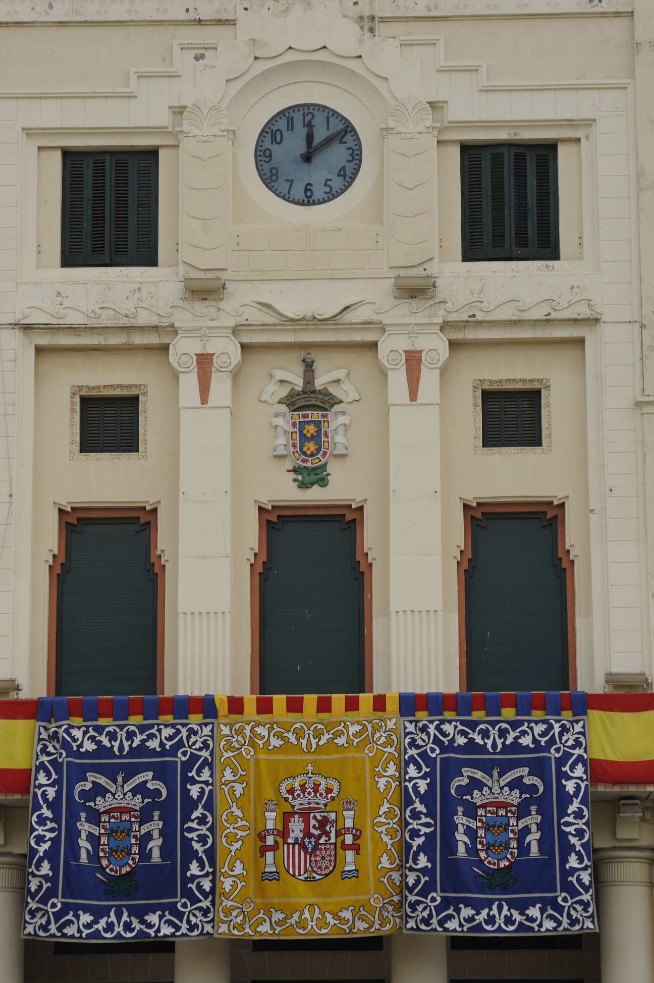 Cream-colored colonial building facade with blue and black shuttered windows, clock, decorative coat of arms, and Spanish heraldic banners hanging below.