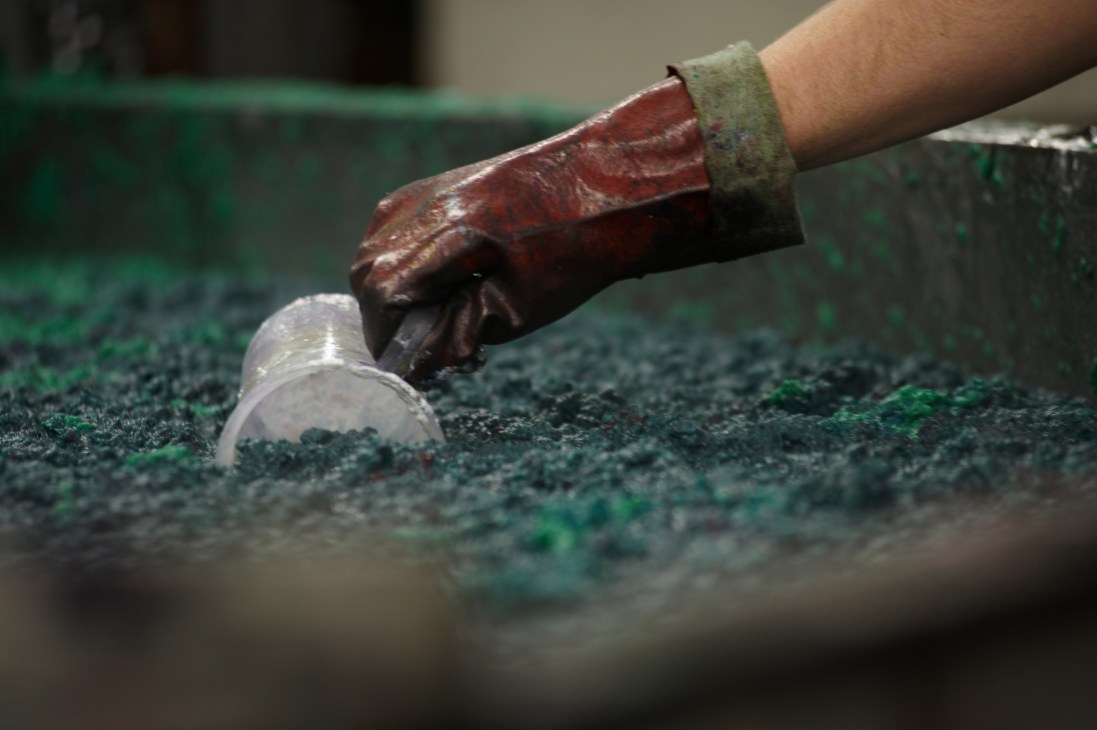 A gloved hand holding wet pulp and dye mixture in a blending chamber at a paper mill.