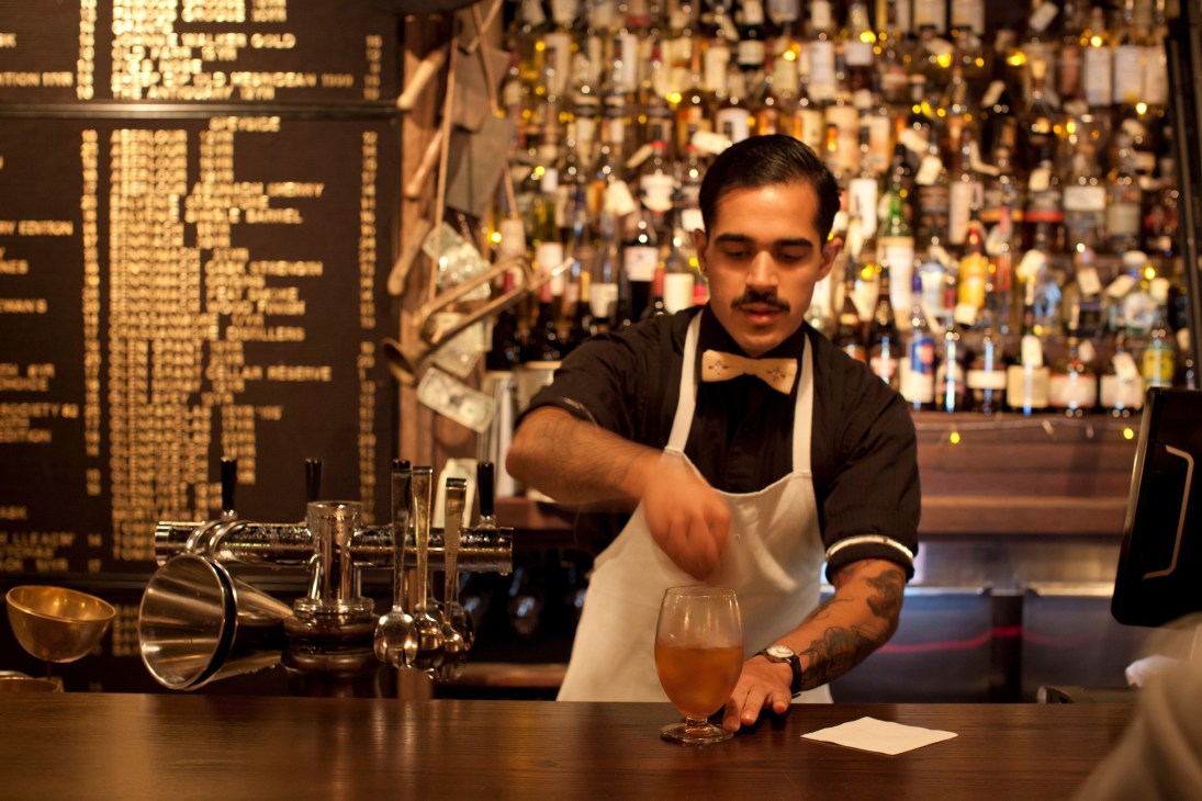 Bartender in white apron and bow tie preparing a cocktail at The Baxter Inn with bottles visible behind the bar.
