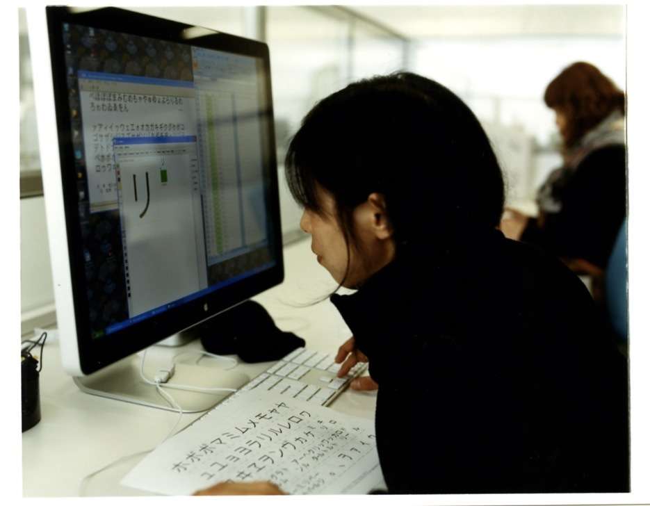 A Morisawa designer works at a computer displaying Japanese characters in font design software while reviewing printed character samples.