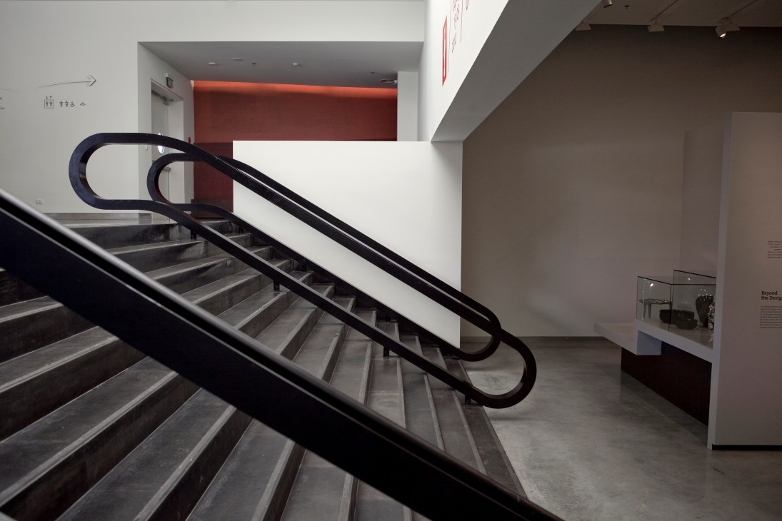 Modern staircase with black handrails leading down through a minimalist gallery space with white walls and red accent lighting.