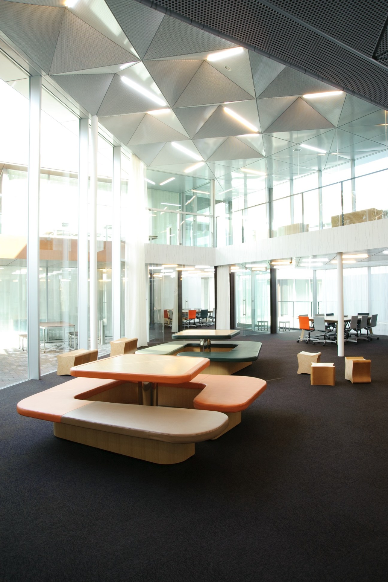 Modern lobby with G-shaped wooden tables, colorful seating units, triangular ceiling with integrated lighting, and glass walls overlooking forest.