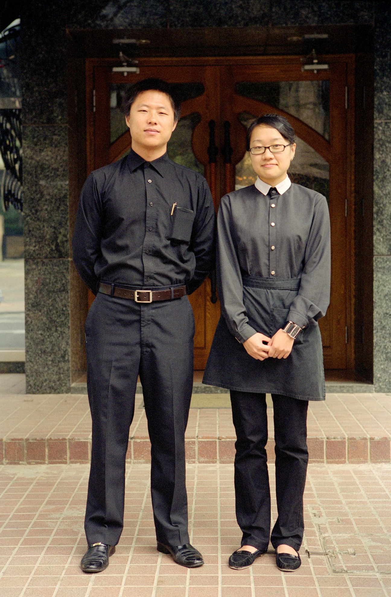 Hugo and Tracy, staff at Riviera Restaurant in Dalian, stand in formal uniforms outside a wooden doorway.