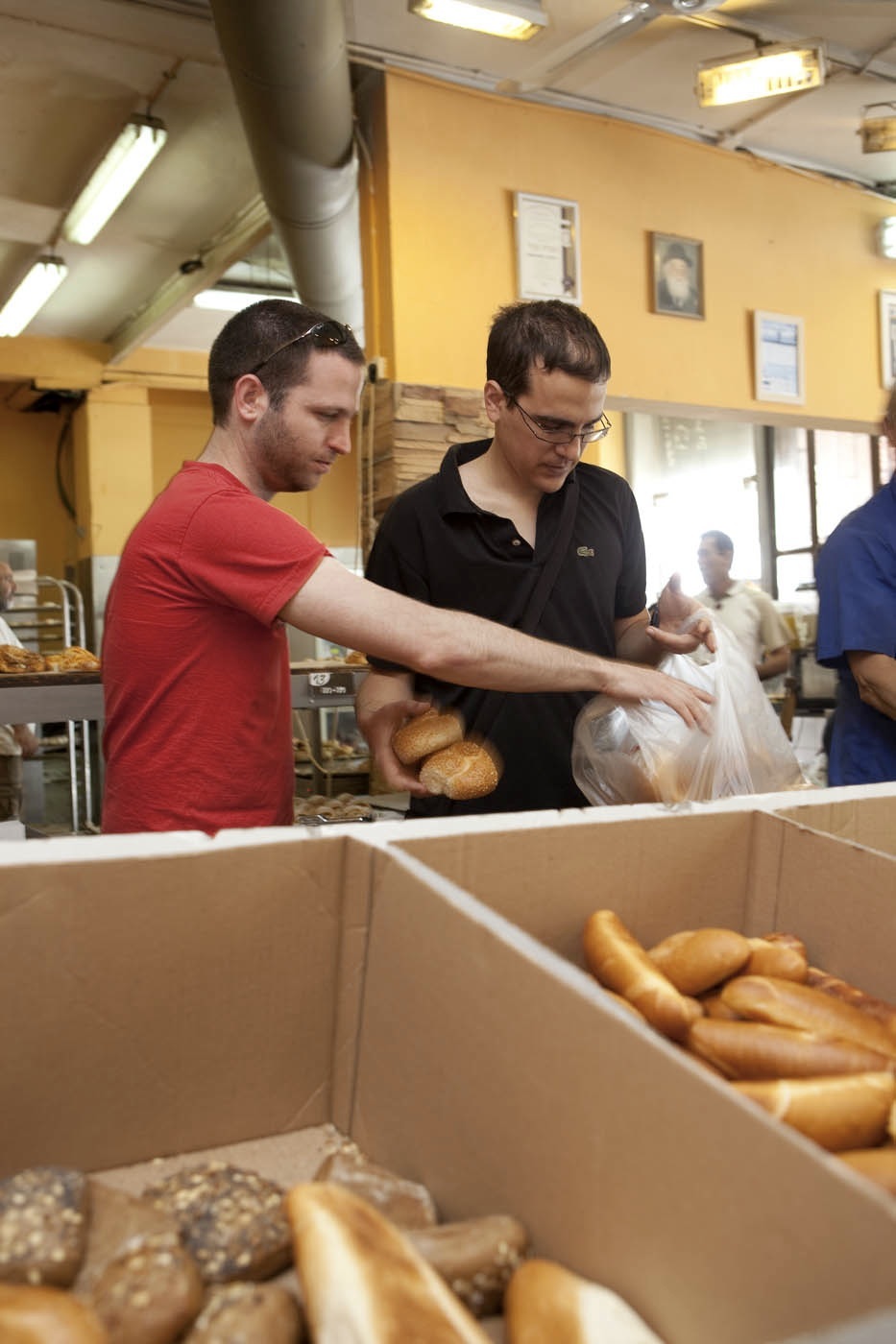 Two men work at a counter in a bakery with yellow walls, packaging fresh bread and baked goods into boxes.