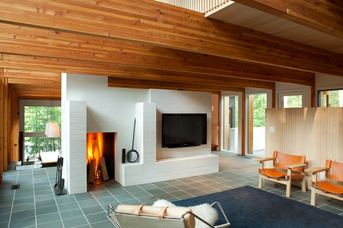 Modern living room with fireplace, wood beams, large windows overlooking forest, and orange leather chairs.