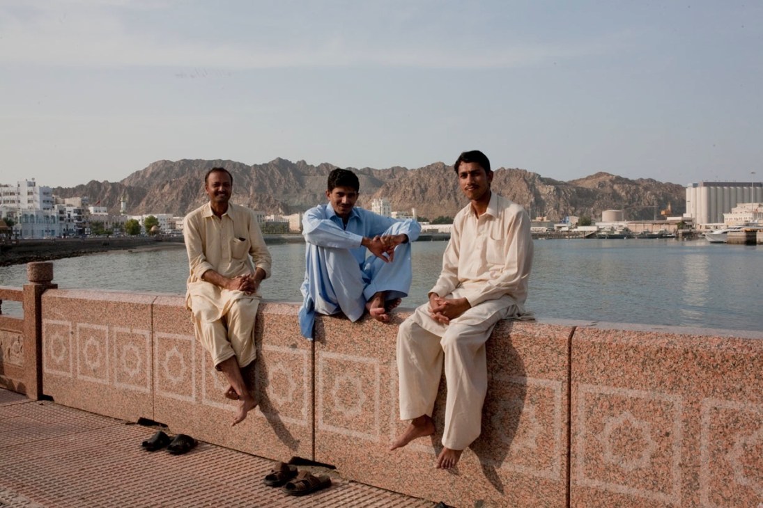 Three men in light-colored traditional dress sit on a waterfront wall with Muscat's mountains and harbor in the background.