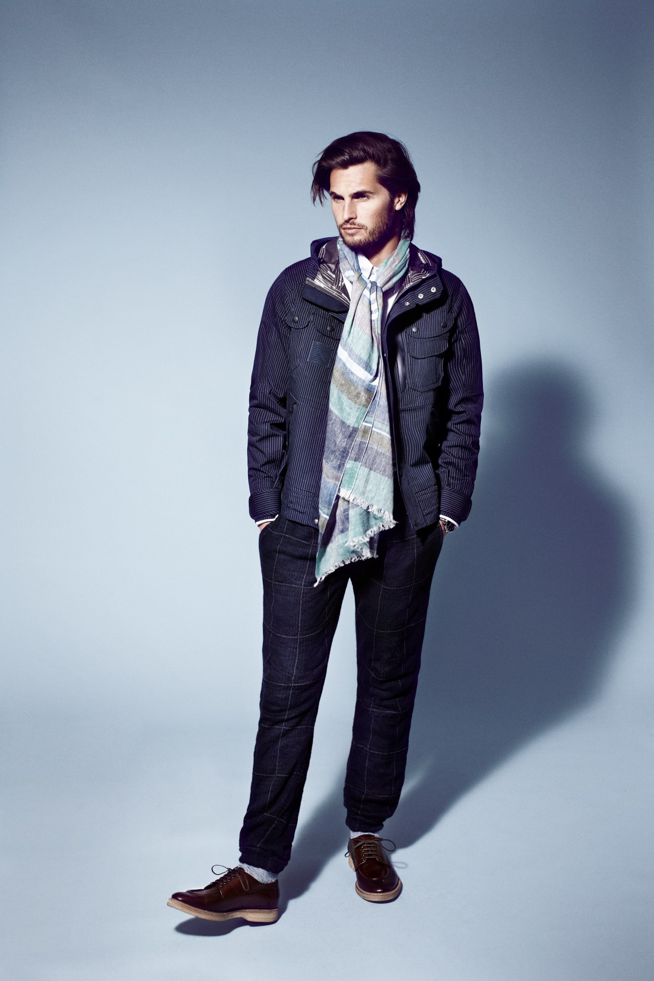 Male model wearing White Mountaineering jacket, Engineered Garments shirt, Bottega Veneta scarf, and Sacai trousers against blue background.