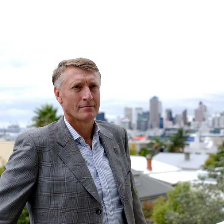 Man in gray blazer and striped shirt standing outdoors with Auckland city skyline in background.