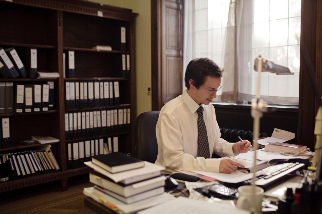 Man in white shirt and tie working at desk surrounded by legal books and files in ornate office.