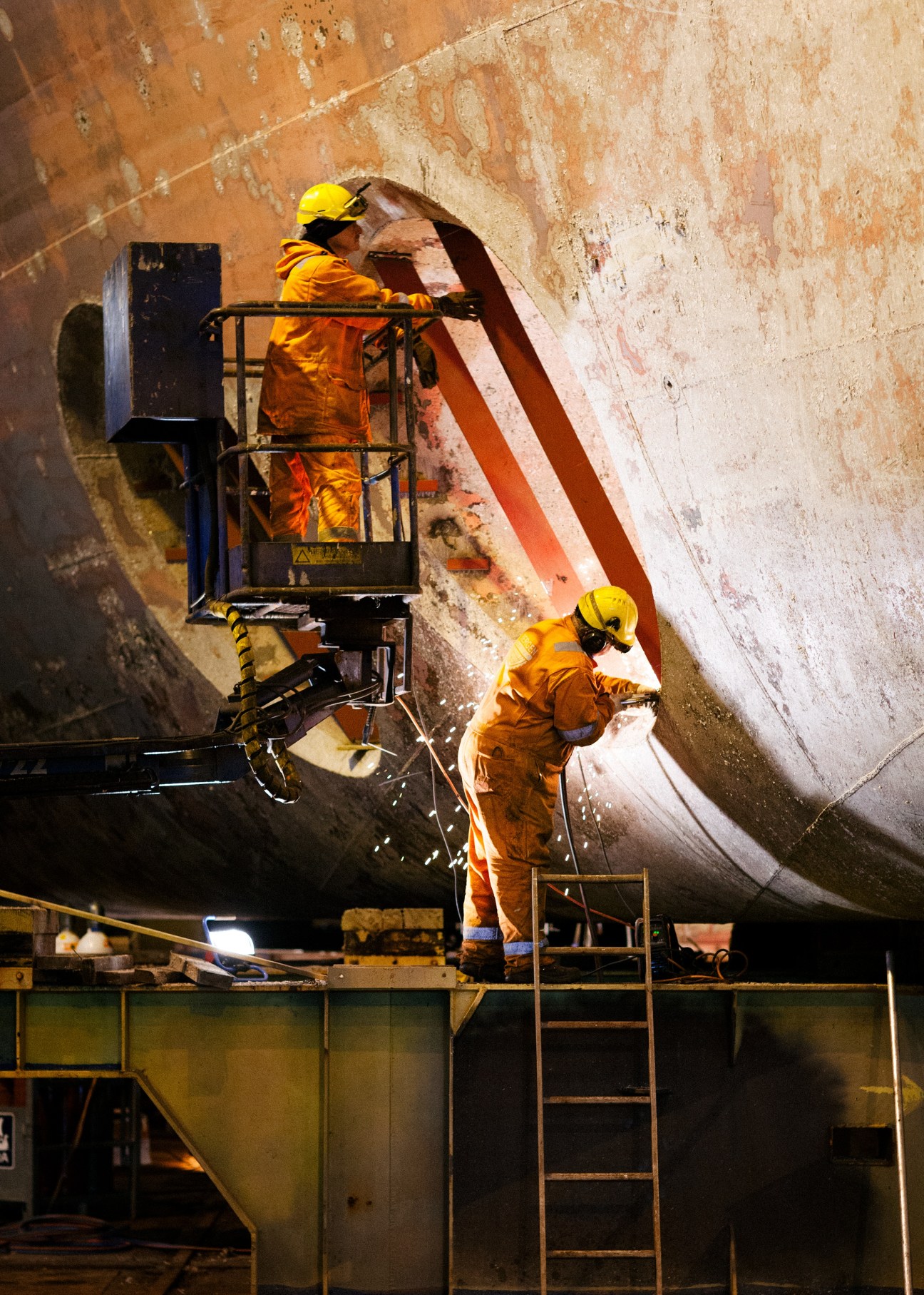 Workers in orange protective gear sandblasting the hull of a large ship at Kimek shipyard in Kirkenes.
