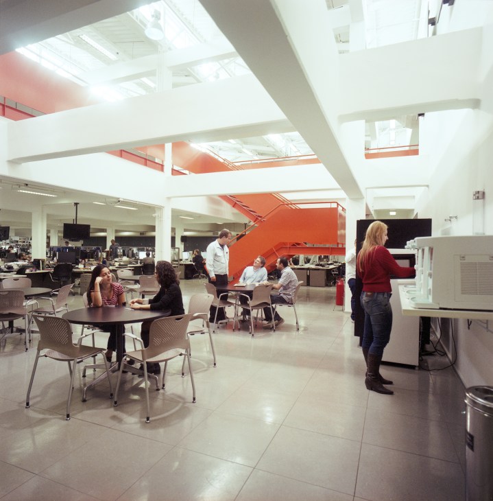 Modern newsroom break area with bright skylights, white columns, red staircase and casual seating where staff relax.