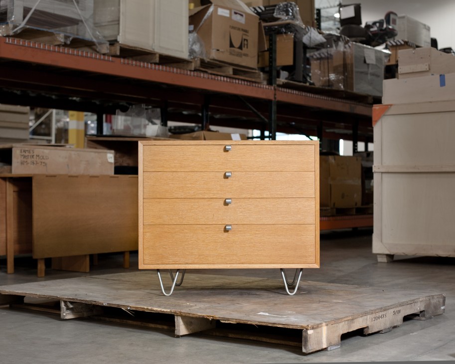 A light wooden four-drawer cabinet with metal handles and hairpin legs displayed in a warehouse.