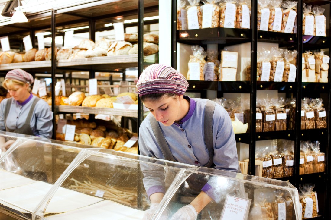 Baker working at counter in Panella bakery with shelves of packaged breads behind.