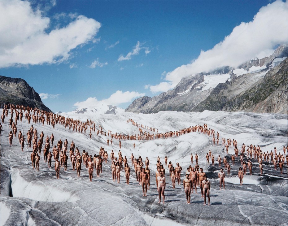 Large group of nude figures standing on a snow-covered glacier surrounded by mountains.