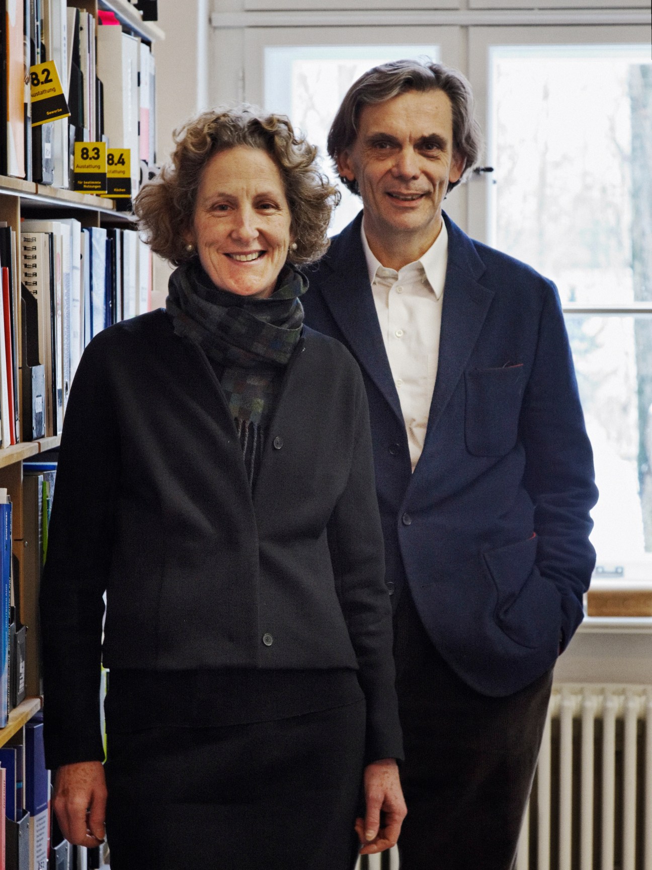 Louisa Hutton and Matthias Sauerbruch stand together in their office next to bookshelves.