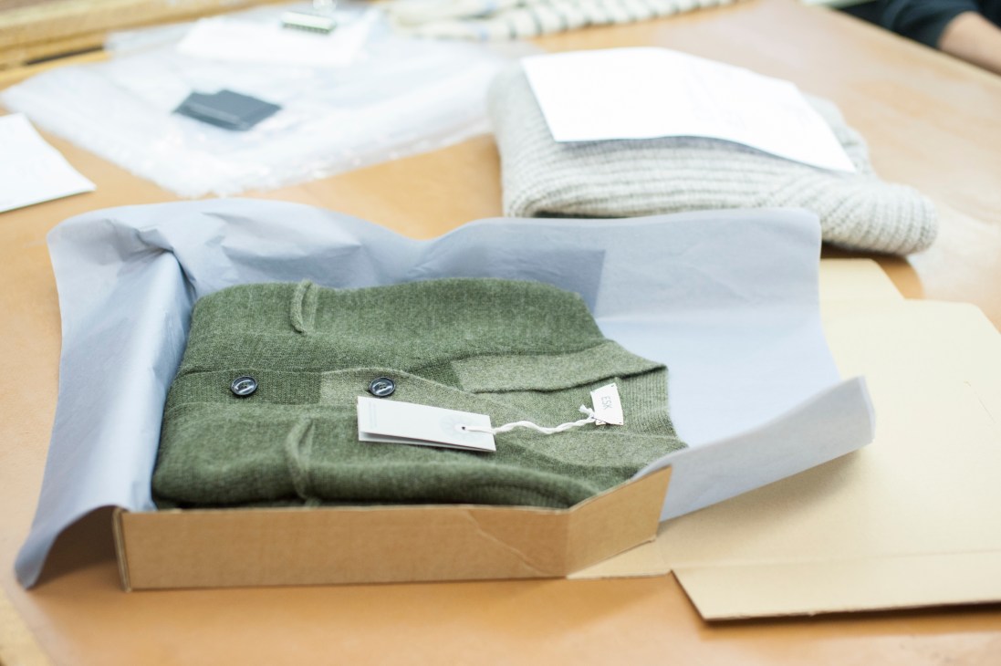 Green knitted cardigan with buttons displayed in a cardboard box with tissue paper on a wooden surface.