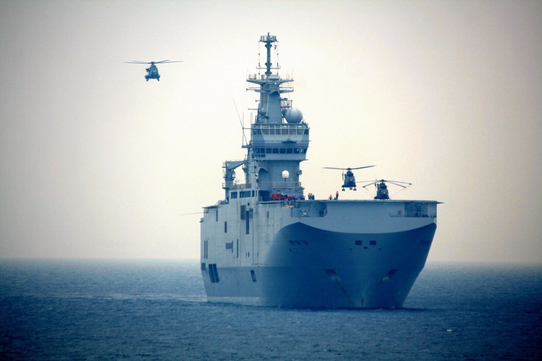 Modern amphibious assault ship at sea with helicopters in flight nearby.