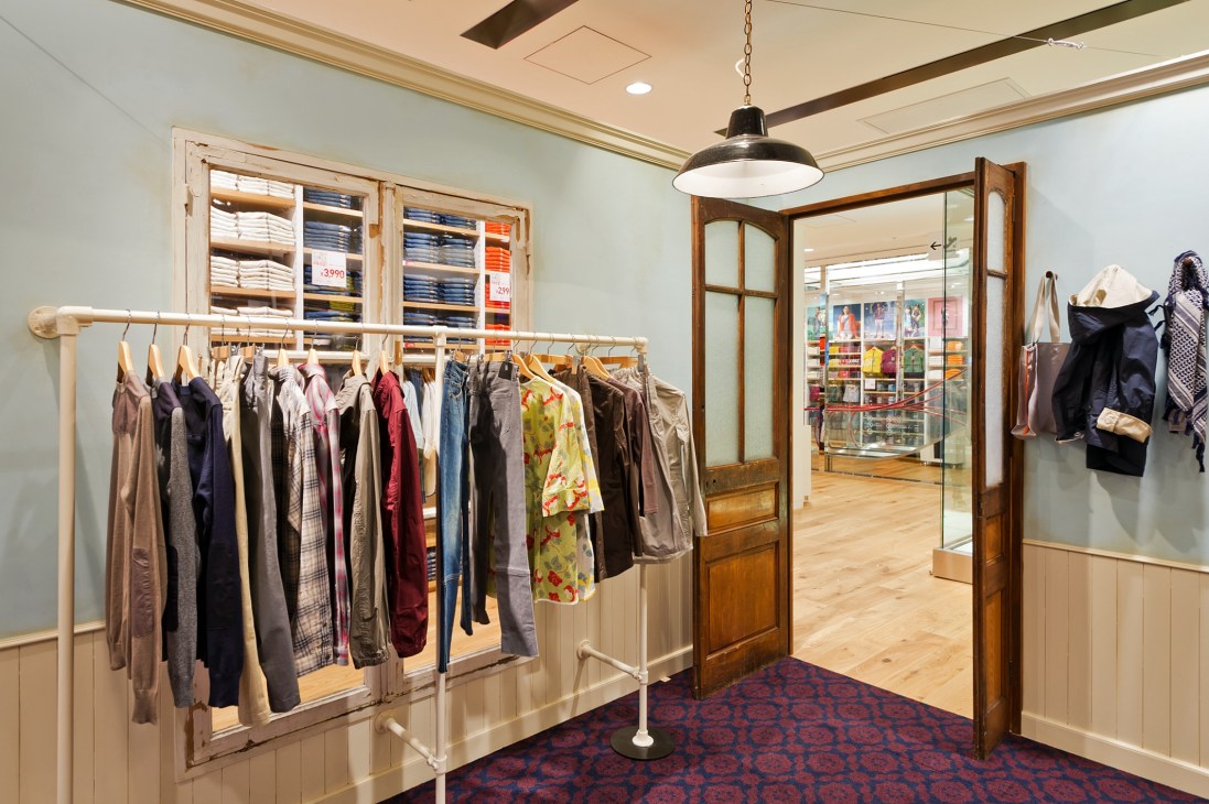 Dover Street Market Ginza interior showing UU collaboration clothing displayed on racks with wooden cupboards and pendant lighting.