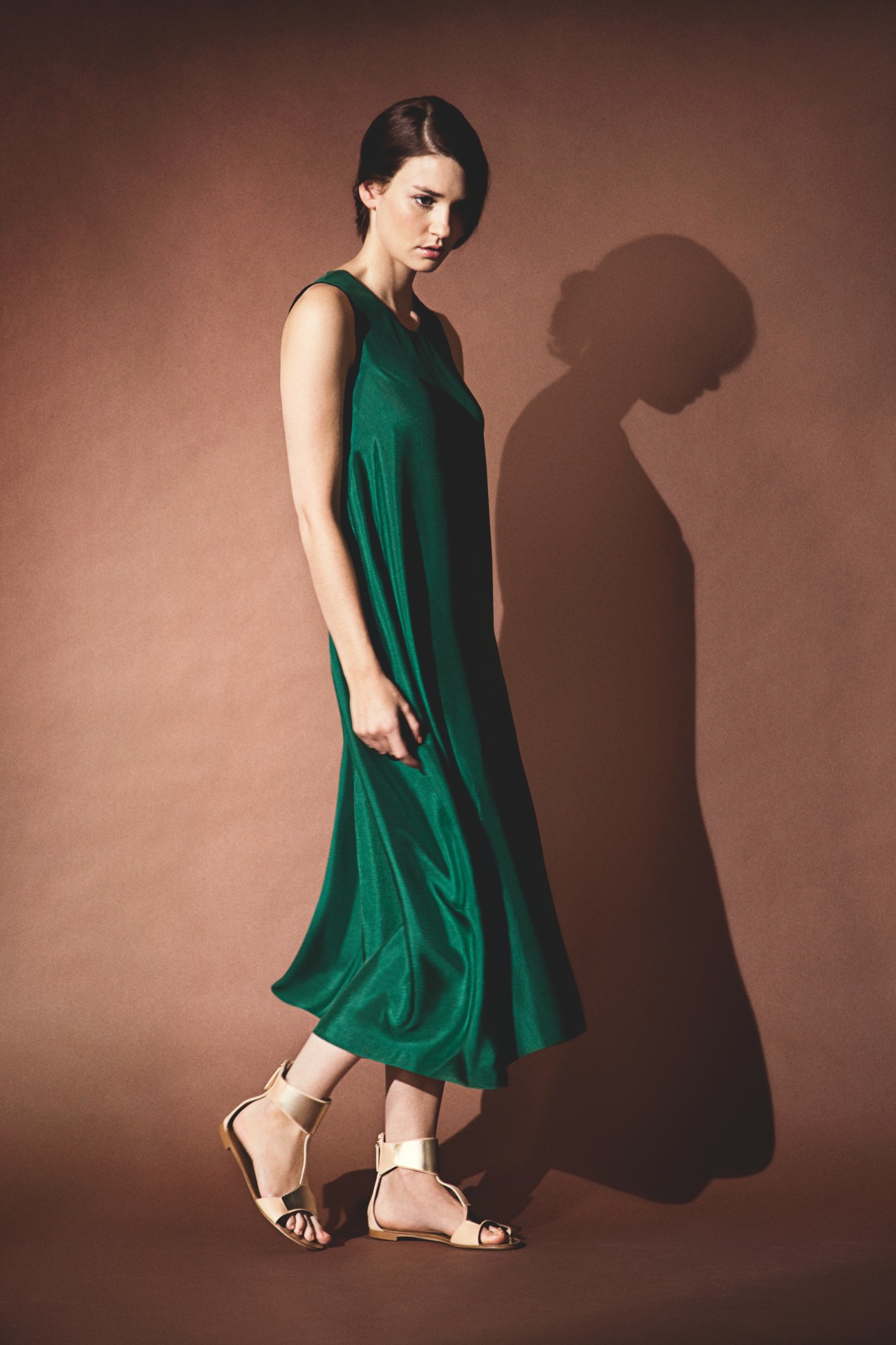 Woman wearing an emerald silk asymmetric dress with loose fit and metallic sandals against a brown background.