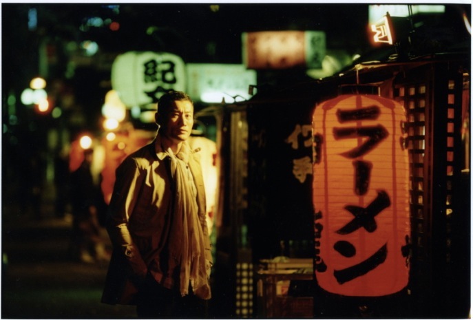 Model wearing Nanamica coat and Comme des Garçons trousers stands on a nighttime Fukuoka street lined with illuminated red lanterns.