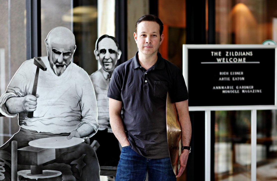 Paul Francis stands next to illustrated figures of cymbal makers at Zildjian's Massachusetts factory entrance.