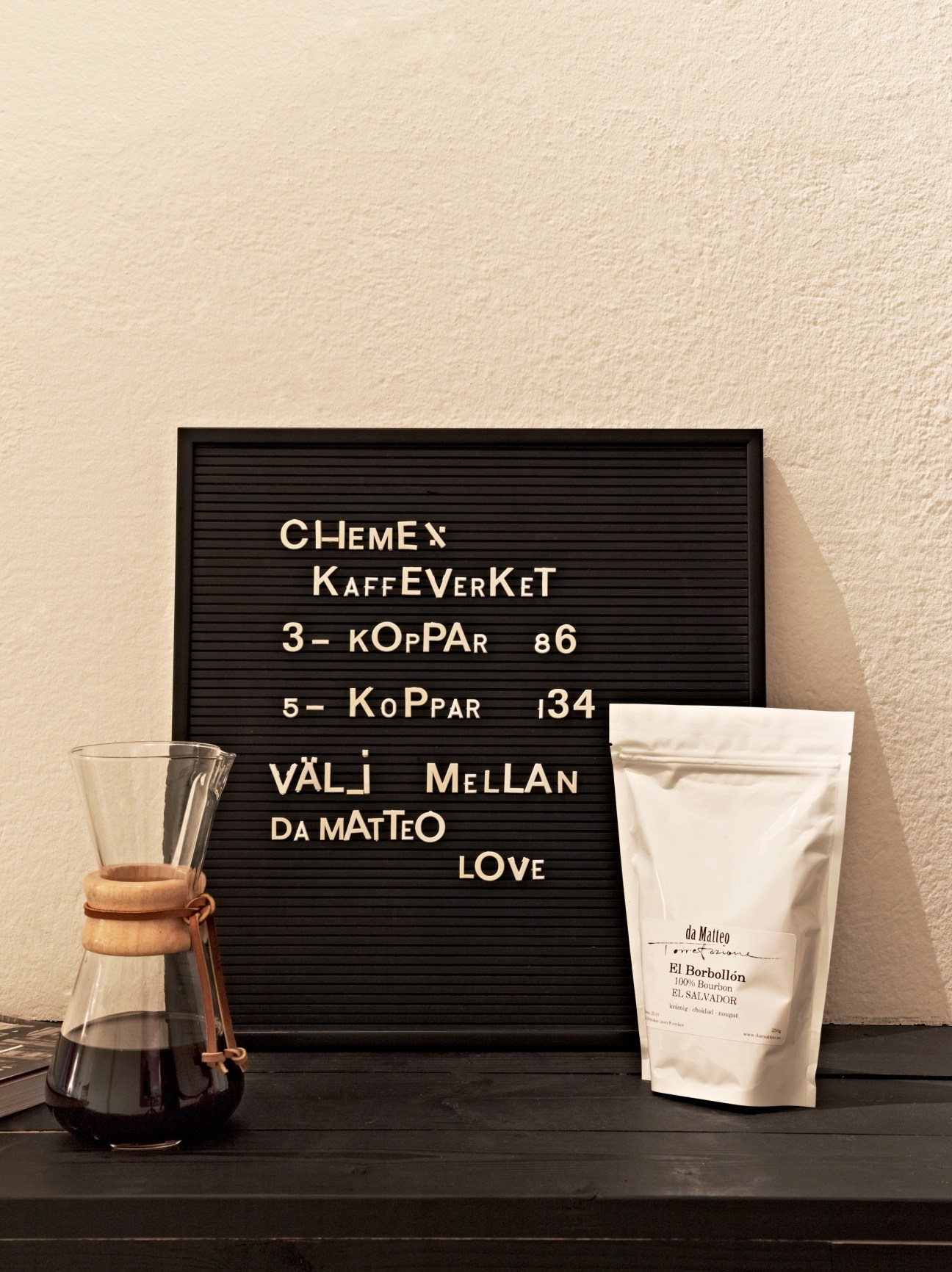 Chemex coffeemaker with wooden collar next to da Matteo coffee beans package and letterboard menu against cream wall.