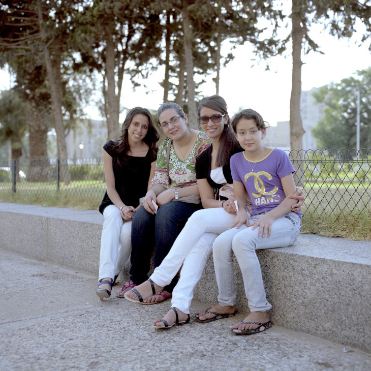 Four young women sitting together on a concrete bench under tall trees along a tree-lined avenue.