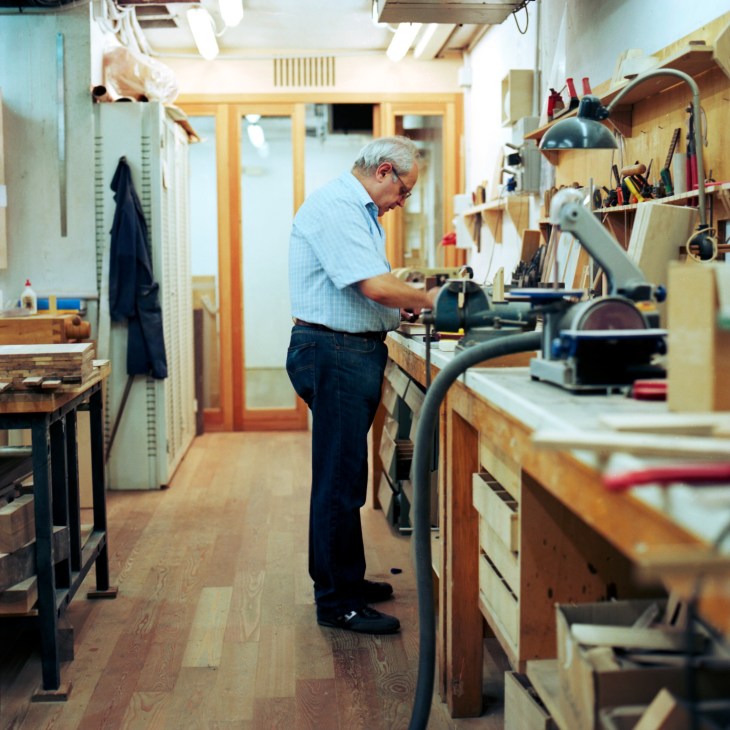 Michele De Lucchi works at a wooden workbench in his Milan model-making workshop surrounded by tools.