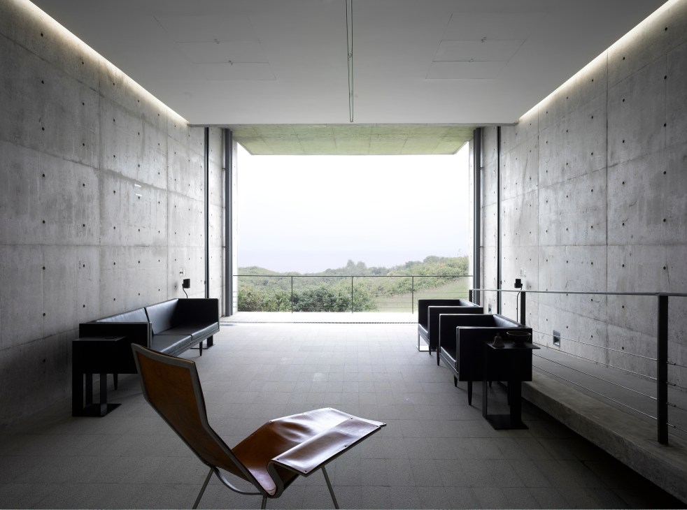 Minimalist concrete interior with sparse furniture overlooking lush tropical landscape through large windows.