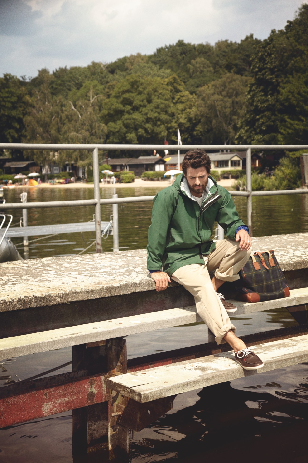 Man in green jacket and cream trousers sitting by a lake with forested hills behind.