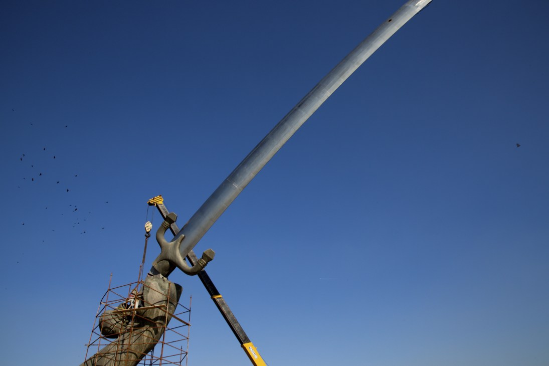 Workers repair a large bronze sword monument with scaffolding under clear blue sky.