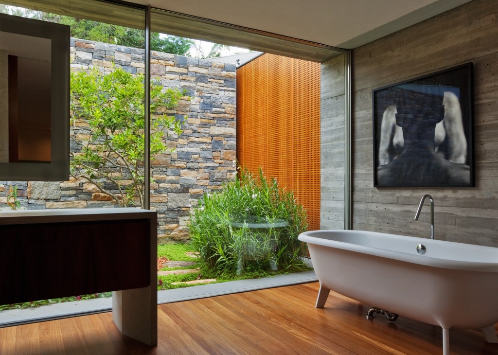 Modern bathroom with freestanding white bathtub, wooden floor, and large glass wall opening to garden with stone wall and plants.