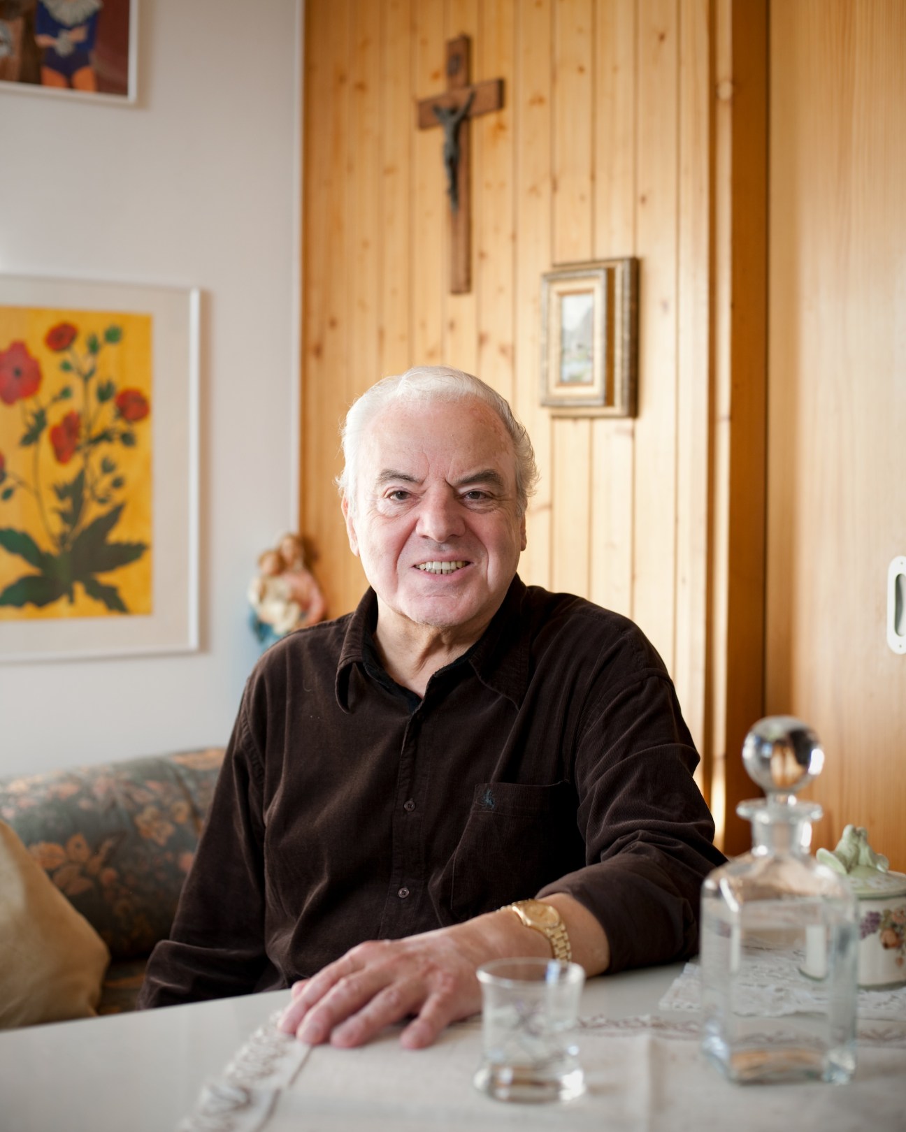 Hans Rubey, singer, smiles at camera in his apartment with artwork and wooden cross on wall.