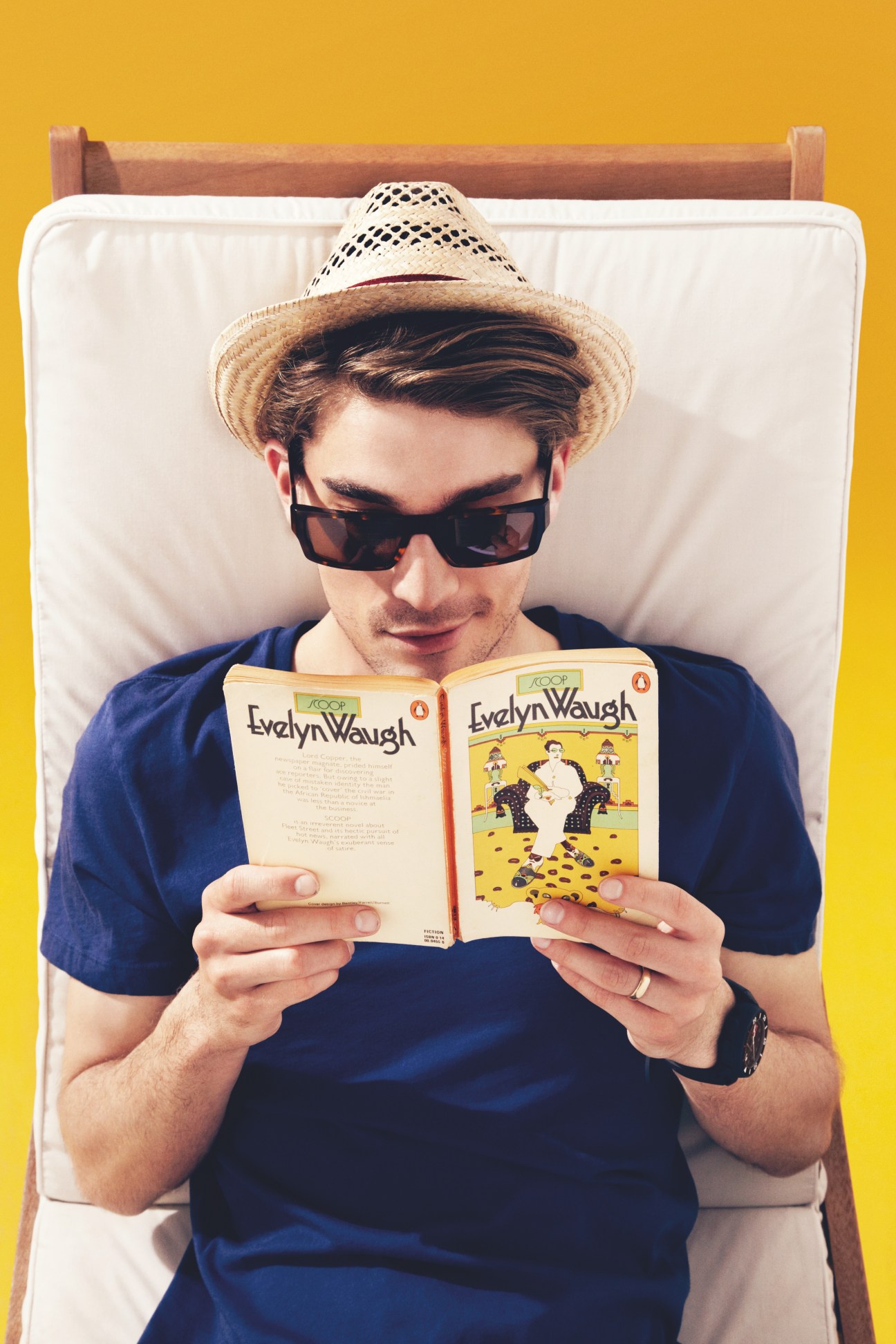 Man lounging on bed wearing sunglasses and hat while reading an Evelyn Waugh book