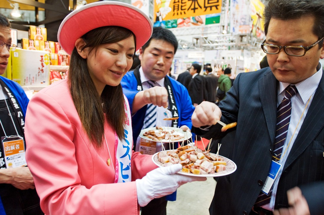 A woman in a pink jacket and red hat serving food samples to attendees at a trade show.