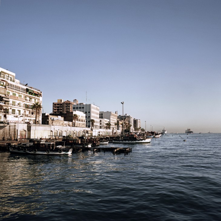 Fishing boats moored along the Suez Canal waterfront in Port Said, with multi-story residential and commercial buildings lining the shore.