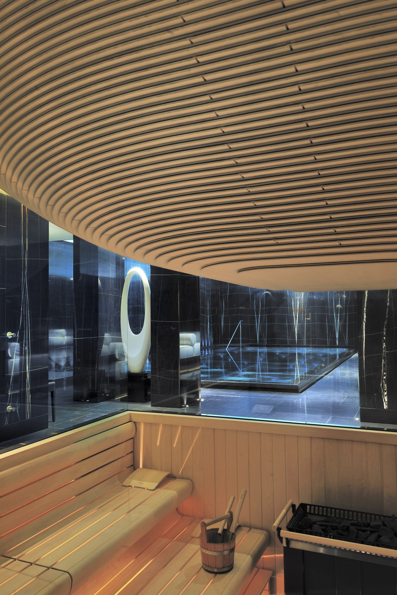 Modern spa sauna with curved wooden ceiling, black marble walls, and illuminated oval light fixture overlooking a swimming pool.