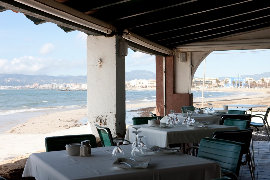 El Bungalow's beachfront terrace with tables set for dining under a concrete pier, overlooking Palma de Mallorca bay.