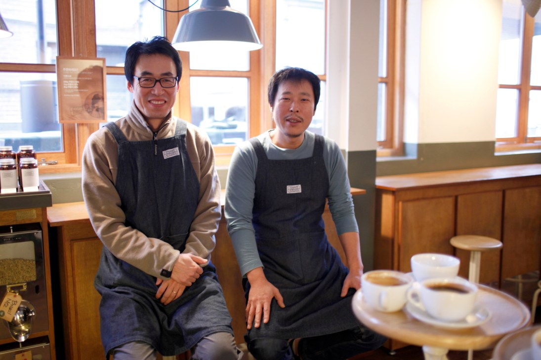 Two male staff members smile at Coffee Kitchen café in Seoul with coffee cups on table.