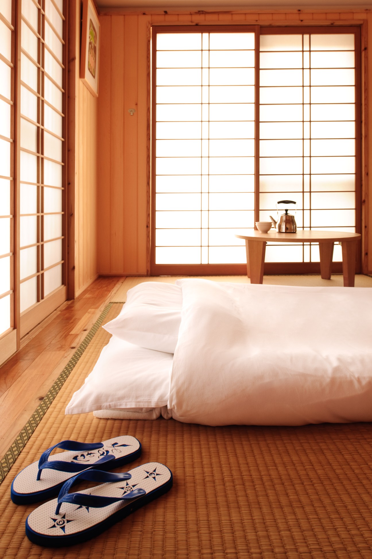 Japanese tatami room with white bedding, wooden furniture, shoji screens, and decorative slippers on woven mat flooring.