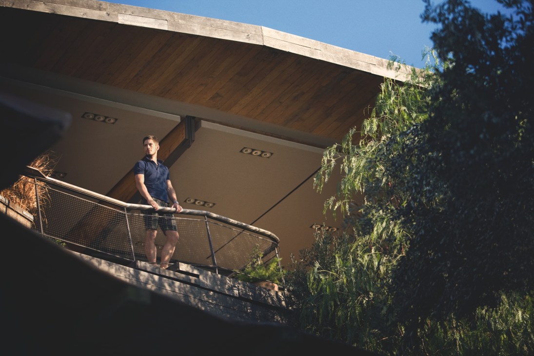 Man wearing Orlebar Brown polo shirt and TS(S) shorts standing on a modern hotel balcony with metal railing and climbing vines.