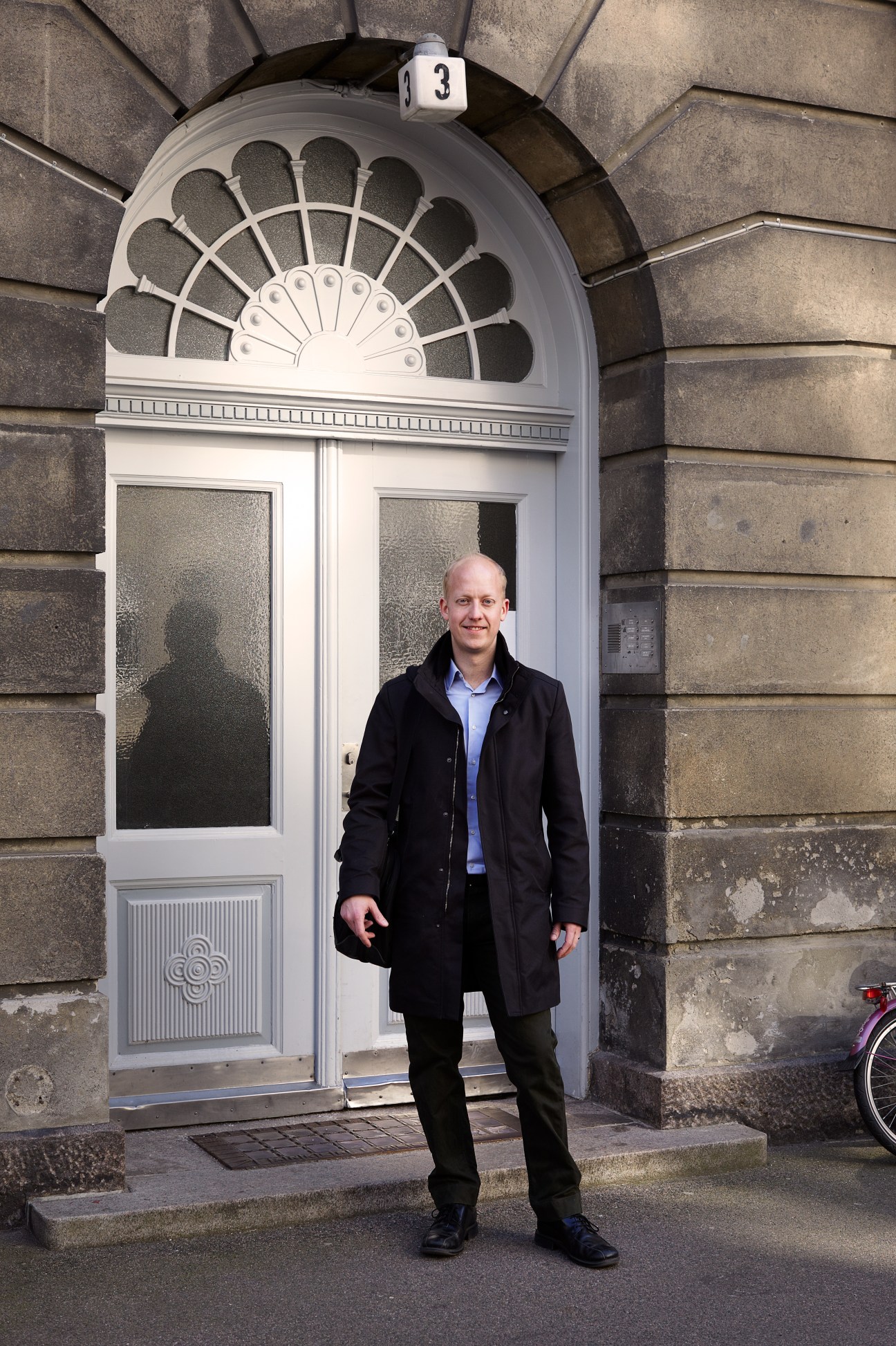 Man in black coat standing in front of a historic Georgian doorway with a fanned transom window.