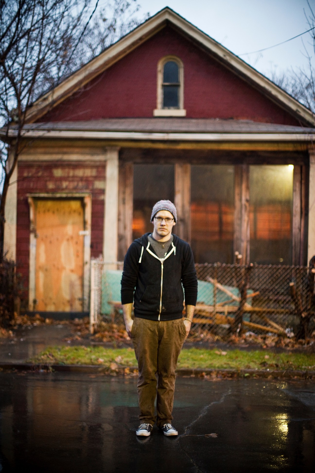 Student Mills Colbert stands in front of a red brick house with a boarded-up door and weathered windows in Buffalo.