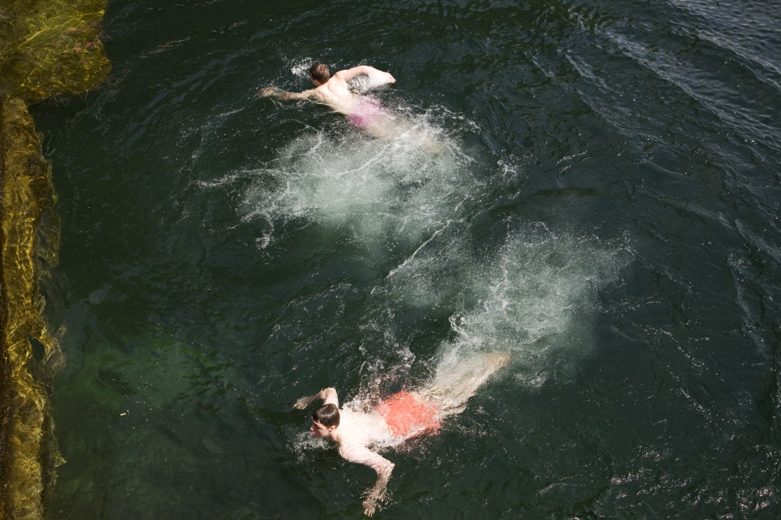 Aerial view of two swimmers in dark green water next to a yellow-green dock.