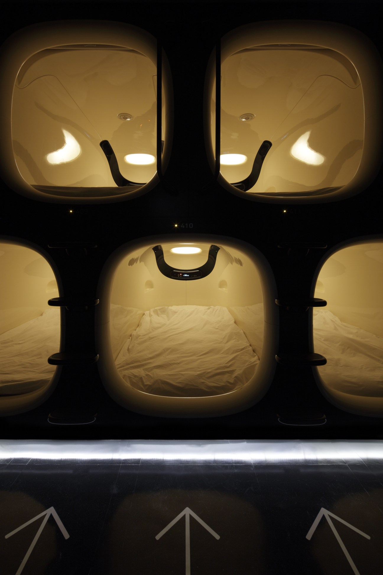 Illuminated capsule hotel sleeping pods with cream-colored interiors and black handles stacked vertically.