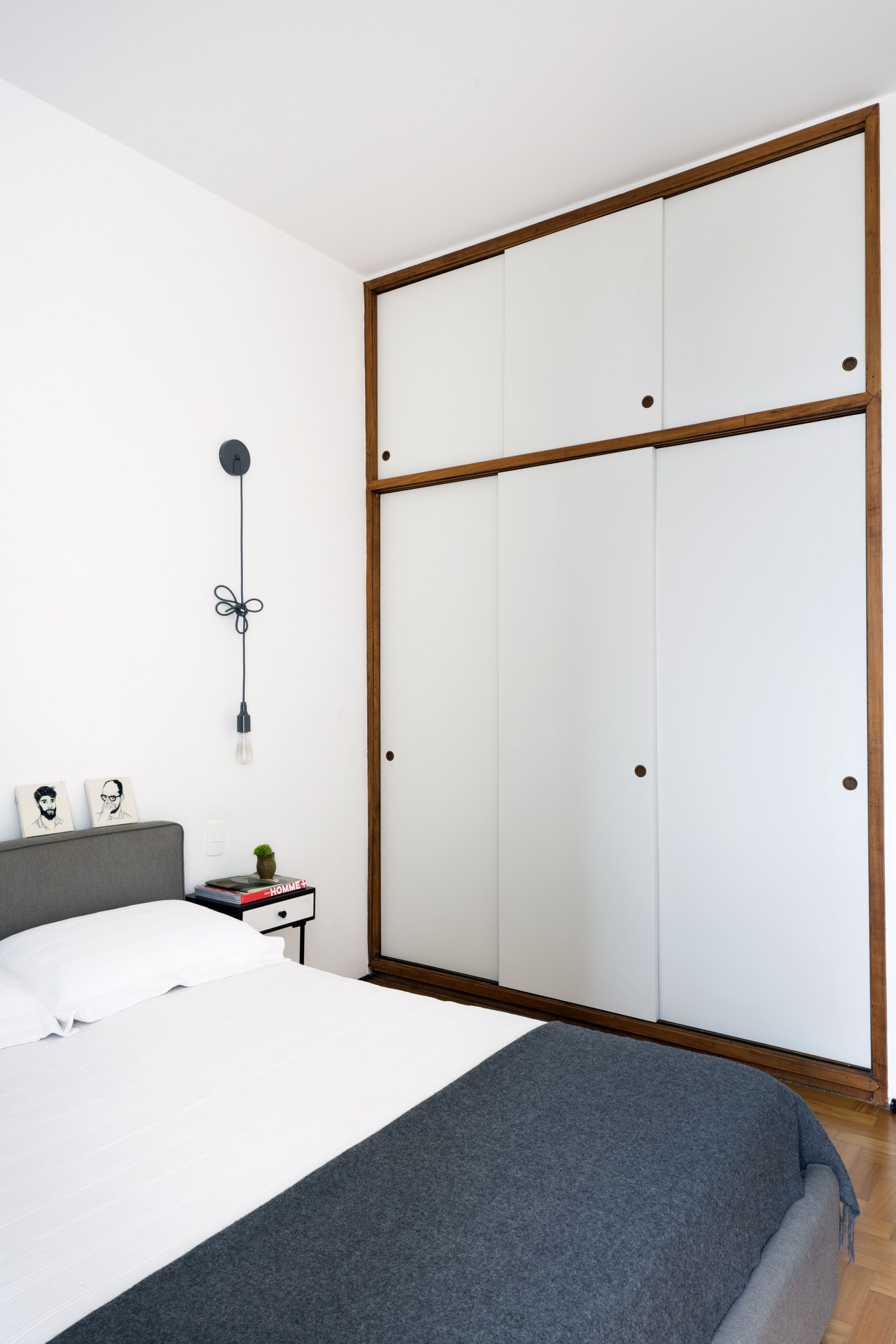 Minimalist bedroom with white walls, wooden wardrobe, gray bed, and pendant light fixture.