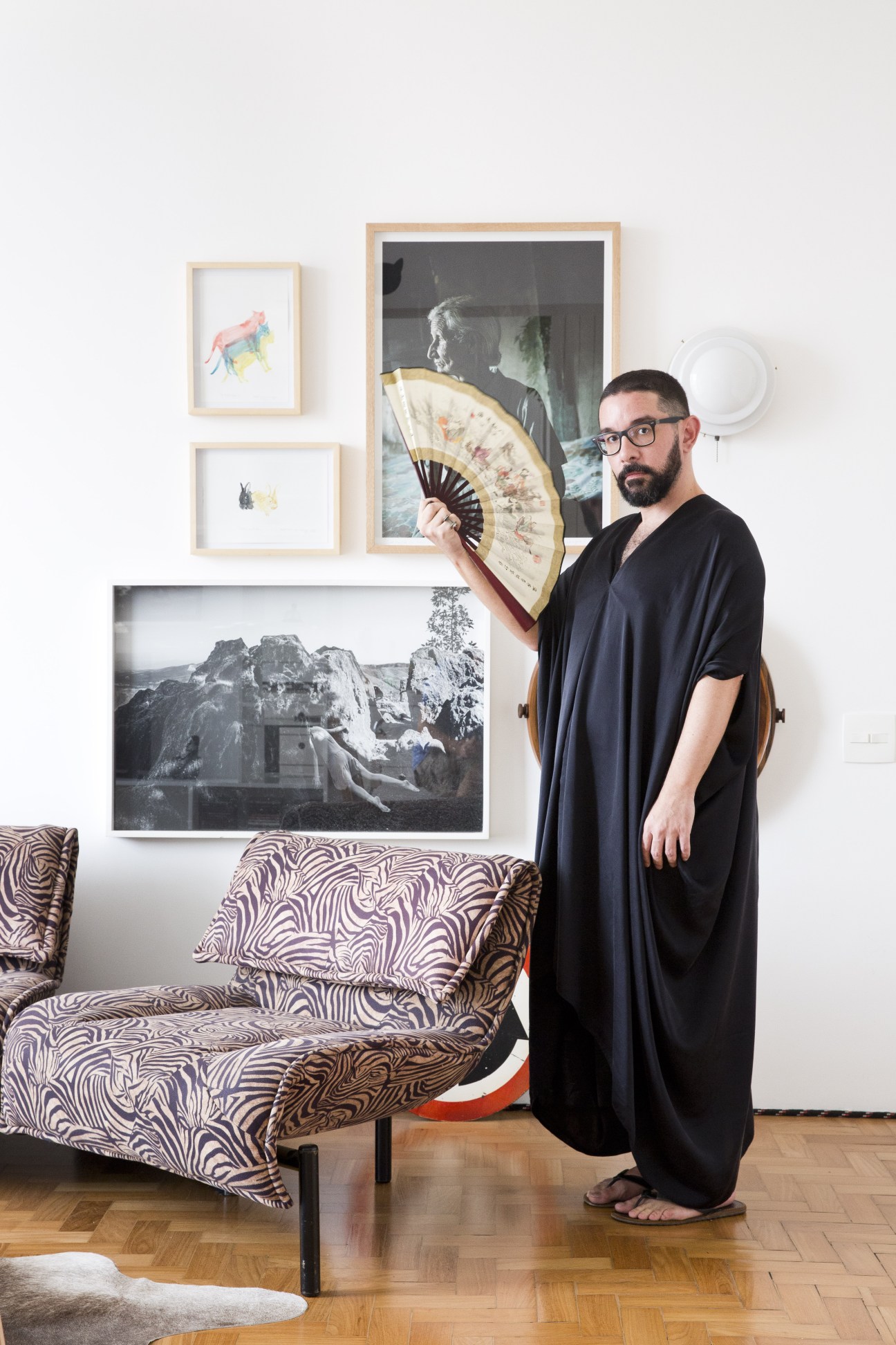 Man in black cardigan holding decorative fan in modern apartment with zebra-print sofa.
