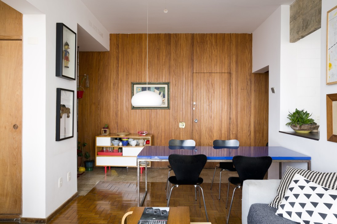 Modern apartment interior with original wood paneling wall, dining table with blue top, black chairs, and potted plant on floating shelf.