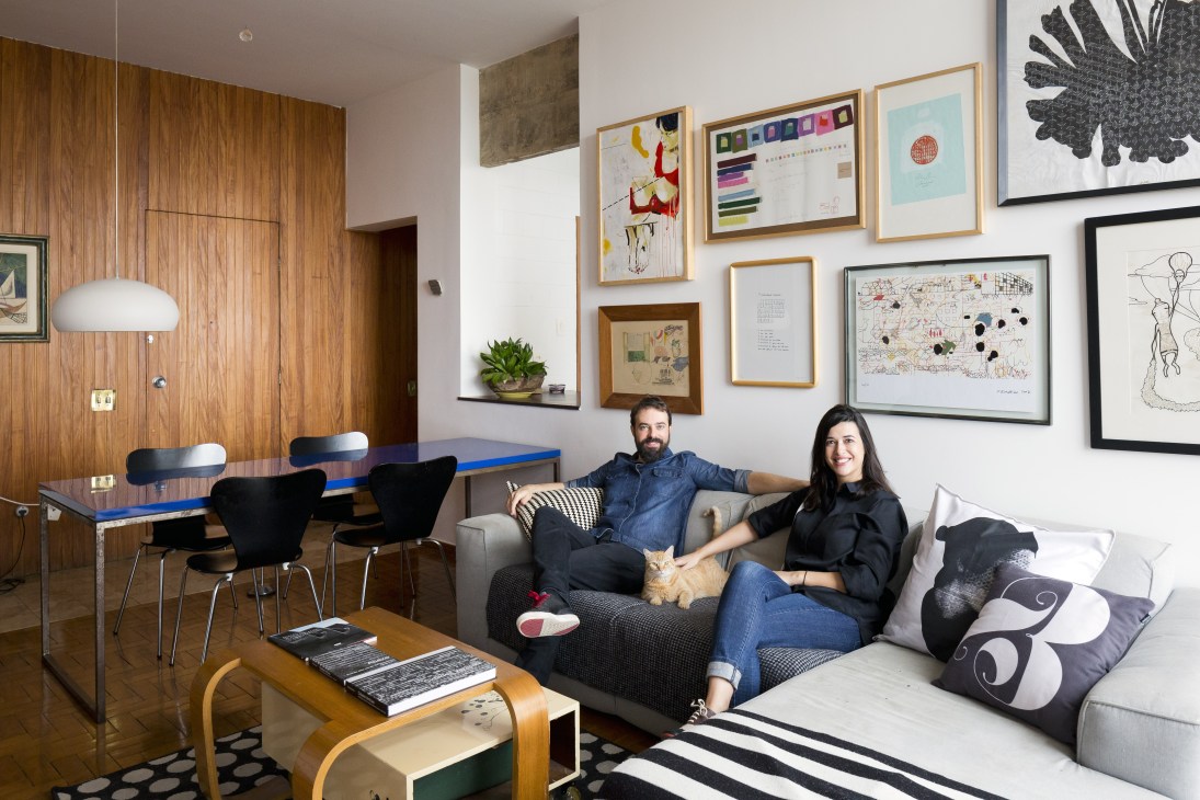 Pedro Morais and Clara Urbano sitting on a gray sofa in their modern JK Building apartment with wood paneling and framed artwork.
